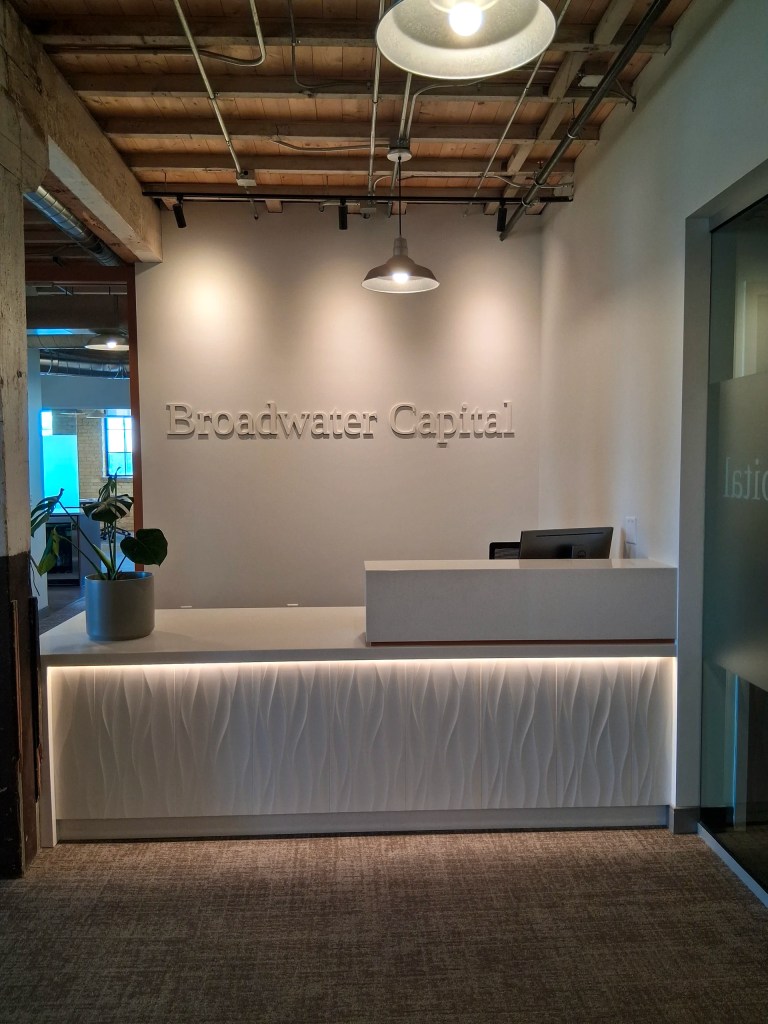 Modern upscale financial office reception area with subtle brushed metal dimensional letter company name sign on a white wall above an illuminated reception desk with exposed beam ceiling