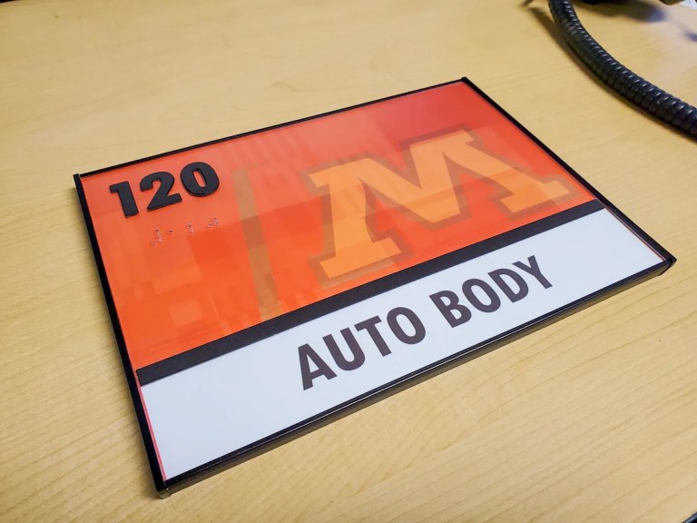 ADA compliant framed room sign with a full-color printed insert featuring a school branded orange and black graphic design, room number 120, Braille, and a nameplate reading Auto Body