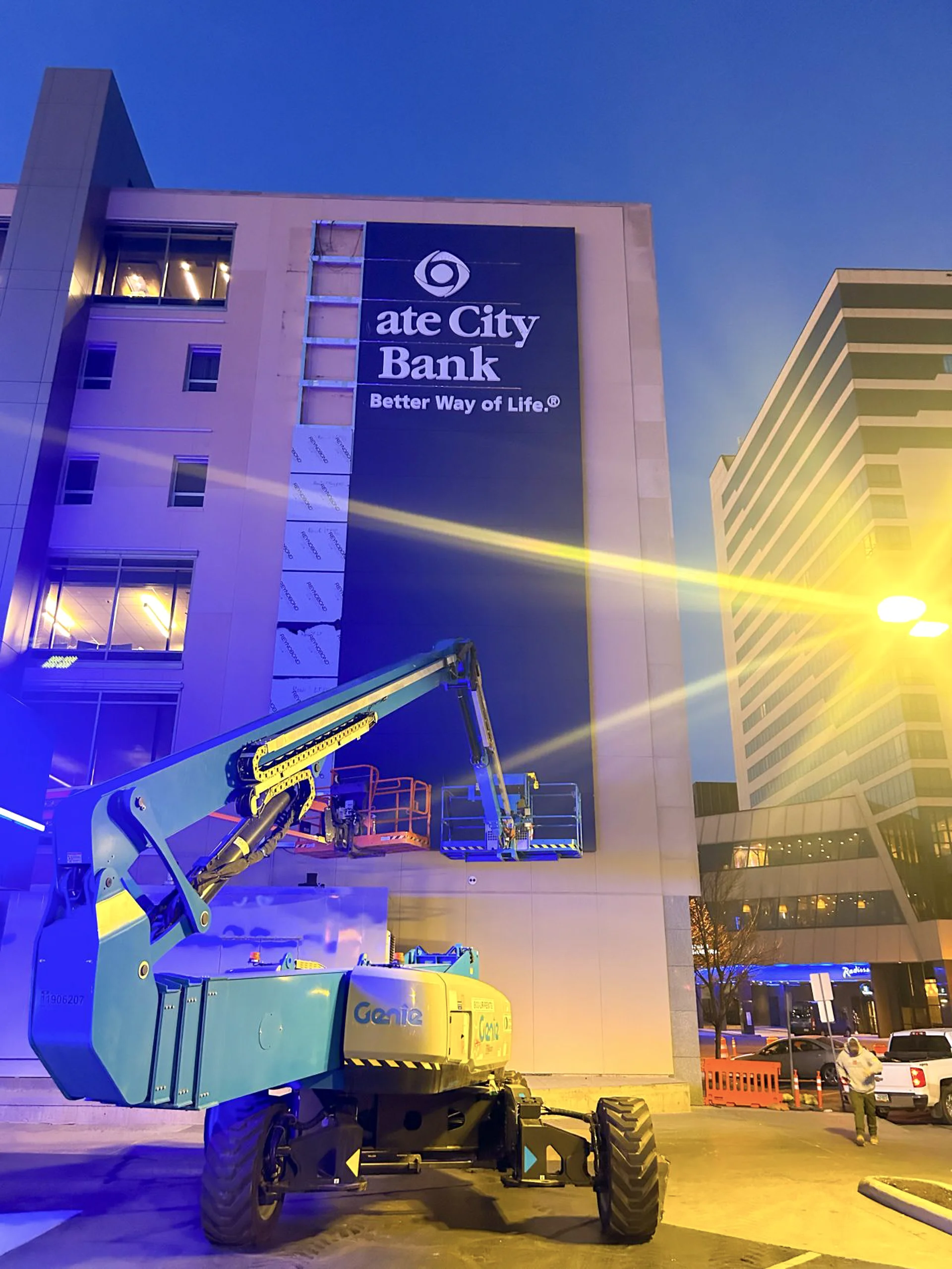 Gate City Bank building sign illuminated at night during installation with Genie boom lift parked in front of the downtown Fargo headquarters